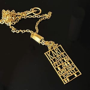 May include: Gold-coloured necklace with a rectangular pendant inscribed with "KARL LAGERFELD" and "MODERNES KUNST". The chain and pendant are made of a shiny, reflective metal, with a geometric design.