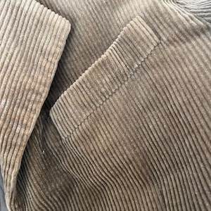 Op de afbeelding: Close-up van een bruine corduroy jas. De jas heeft een getextureerde, geribbelde stof en een knoopsluiting. De afbeelding benadrukt de details van de stof en de afwerking van het kledingstuk.