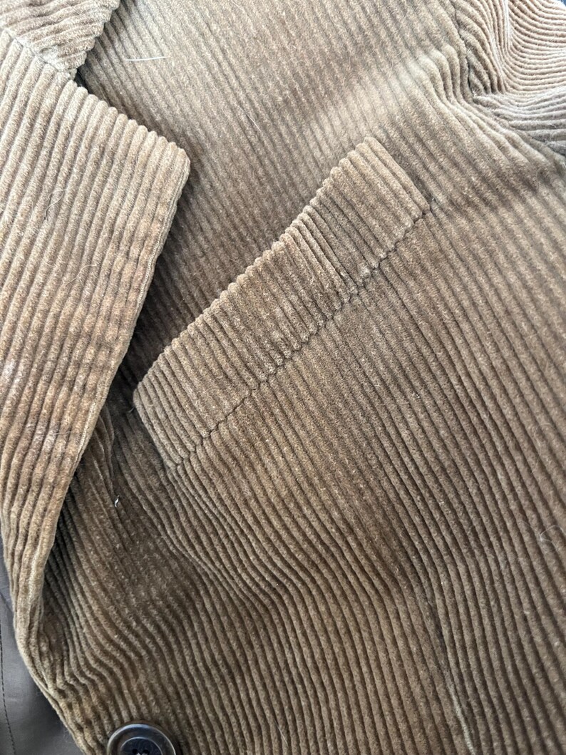 Op de afbeelding: Close-up van een bruine corduroy jas. De jas heeft een getextureerde, geribbelde stof en een knoopsluiting. De afbeelding benadrukt de details van de stof en de afwerking van het kledingstuk.