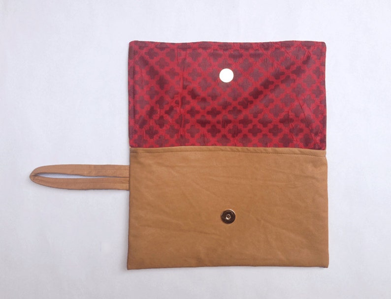 Puede incluir: Un clutch de cuero marr&oacute;n con un forro de tela roja y un cierre magn&eacute;tico. El clutch tiene una sola correa.