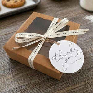 May include: A brown cardboard box with a clear window, tied with a cream ribbon and a "Thank You" tag. The box is on a wooden surface, with a baking sheet of cookies in the background.