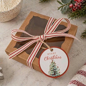 May include: A square, brown gift box with a clear window, tied with a red and white striped ribbon. A round tag with "Merry Christmas" and a Christmas tree illustration hangs from the ribbon. The box is on a light-colored surface.