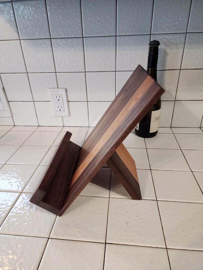 May include: A wooden cookbook stand with a walnut and maple finish. The stand is angled to display a cookbook.