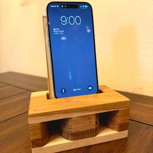 May include: A wooden phone stand with a cutout for a smartphone. The stand has a small speaker hole on each side. A smartphone is in the stand, displaying the time as 9:00. The phone screen shows a weather app with a partly cloudy icon and the temperature 34 degrees.