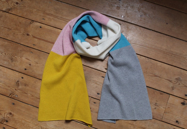 Peut inclure: Une &eacute;charpe ray&eacute;e de couleur jaune, rose, blanc, bleu et gris. L'&eacute;charpe est pli&eacute;e en deux et pos&eacute;e &agrave; plat sur une surface en bois.