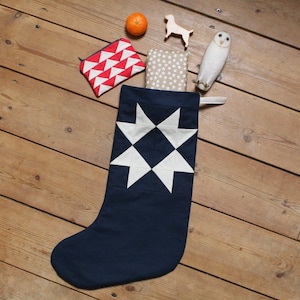May include: A navy blue Christmas stocking with a white star patchwork design. The stocking is filled with small gifts, including a red and white geometric patterned pouch, a wooden dog figurine, and a white owl ornament.