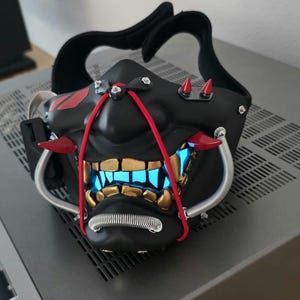 May include: A black face mask featuring a stylized design with gold-colored teeth, red accents, and silver-toned coiled tubing. The mask includes two small red spikes on the upper section and emits a bright blue light from the mouth area.