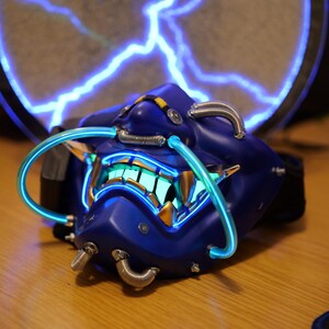 May include: A futuristic, blue Oni mask with glowing neon accents. The mask features a detailed design with metallic tubes and a glowing mouth. The background includes a circular element with a blue lightning design.