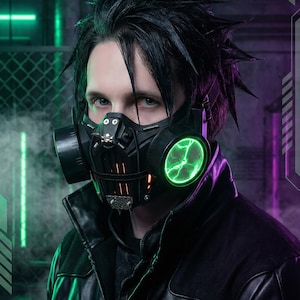 Peut inclure: Un masque à gaz noir avec des détails verts lumineux et un emblème de crâne. La personne porte une veste en cuir noire. L'arrière-plan présente un éclairage néon vert et violet.