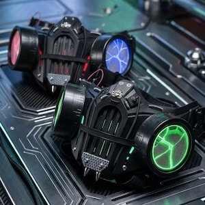 May include: Two black respirator masks featuring a central grill, metallic spikes at the chin, and circular side filters. One mask displays glowing green cracked patterns, while the other features red and blue illuminated filter designs.