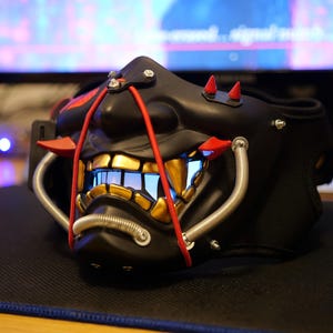 Può includere: Maschera Oni nera con denti dorati, dettagli rossi e tubi argentati. La maschera ha un'espressione minacciosa con una luce blu che emana dalla bocca. Accessorio cosplay o costume.