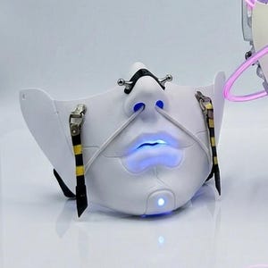 Op de afbeelding: Een witte, futuristische masker met een gloeiende blauwe mond en neus. Het masker heeft zwarte en gele accenten en een uniek, gestileerd ontwerp. Het masker wordt verlicht met blauw licht, wat een opvallend visueel effect creëert.