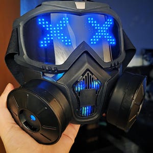 May include: Black and gray futuristic mask with blue LED lights forming an "X" shape on the lenses. The mask has two circular filters on the sides and a strap for securing it. The overall design is sleek and modern.
