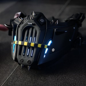 Puede incluir: Una máscara negra de estilo futurista con púas plateadas y una correa amarilla y negra. La máscara tiene un diseño de rejilla y luces LED azules. La máscara está hecha de un material duro y duradero.