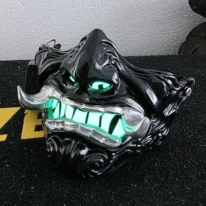 May include: A black mask with glowing green eyes and teeth, featuring silver detailing. The mask has an intricate, textured design. Wires are visible at the back.