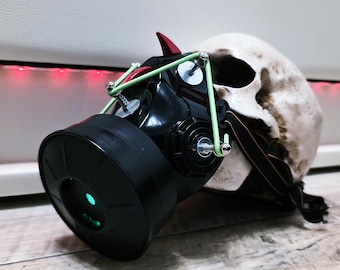May include: A black gas mask featuring two red horn-like protrusions on the top, secured with light green elastic cords and silver hardware. The mask is positioned over a realistic human skull model, with a large circular filter attached to the front.