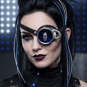 May include: A black monocle featuring a silver skull emblem centered within a deep blue circular lens, attached to a black elastic strap. The monocle includes a metallic side detail with a coiled spring component and thin blue fiber optic strands.