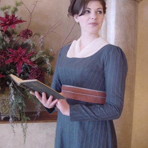 May include: A woman in a blue dress with a white collar and a brown belt is holding a book. She is standing in front of a white pillar.