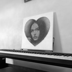 May include: A black and white string art portrait of a woman's face within a heart shape. The artwork is displayed on a white canvas, resting on a black piano. A framed picture hangs on the wall above the piano.
