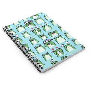 Puede incluir: Cuaderno de espiral con una cubierta azul claro con un patrón repetido de ranas verdes de dibujos animados con varias expresiones. Las ranas tienen vientres blancos y ojos negros. Algunas ranas tienen corazones.