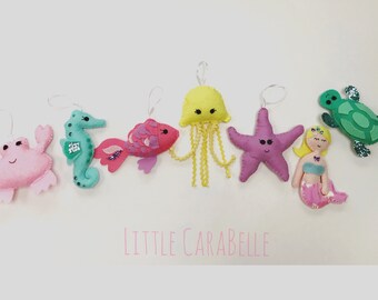 May include: A set of seven felt sea creature ornaments, including a pink crab, a teal seahorse, a pink and purple fish, a yellow jellyfish, a purple starfish, a pink mermaid, and a green turtle. The ornaments are all handmade and have embroidered details. The text "LITTLE CARABELLE" is printed below the ornaments.