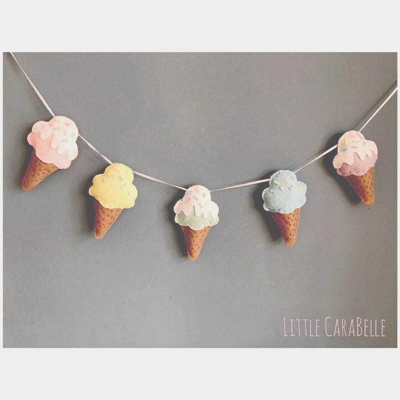 May include: A garland of five felt ice cream cones in pink, yellow, green, blue, and pink with sprinkles. The cones are strung together with white string and the garland says "LITTLE CARABELLE" at the bottom.