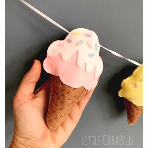 May include: A felt ice cream cone garland with pink and yellow ice cream cones with white frosting and colorful sprinkles. The garland is strung on a white ribbon.