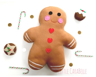 May include: A brown felt gingerbread man with red heart decorations and pink cheeks, alongside candy canes, a Christmas pudding, and a star-shaped pastry. The gingerbread man has white icing details and black eyes. Gold confetti on a white background.