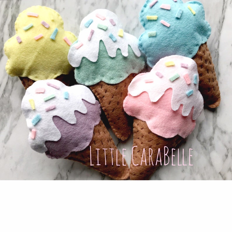 May include: Five felt ice cream cones with different colored ice cream and sprinkles. The ice cream flavors are yellow, green, purple, pink, and blue. The cones are brown. The text "LITTLE CARABELLE" is visible in the bottom right corner of the image.