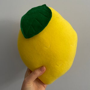 Puede incluir: Un limón de peluche amarillo con una hoja de fieltro verde en la parte superior. El limón es aproximadamente del tamaño de la mano de una persona.