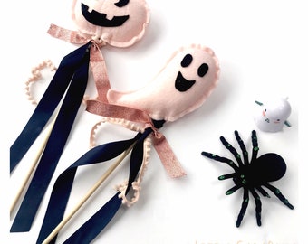 Può includere: Due bacchette di Halloween in feltro rosa con volti di fantasma e zucca in feltro nero. Le bacchette hanno un nastro nero e un nastro color pesca con un bordo di pon pon bianco. Un ragno nero con accenti verdi su uno sfondo bianco. Il testo "LITTLE CARABELLE" è nella parte inferiore dell'immagine.