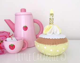 May include: A pink teapot, teacup, and a small box with a tea bag. A yellow felt cupcake with white frosting and a gold glitter candle sits on a white surface. The text "LITTLE CARABELLE" is visible in the bottom right corner of the image.