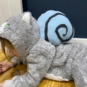 Puede incluir: Un niño con un disfraz de gato gris y blanco peludo con una concha de caracol azul y negra en la espalda.