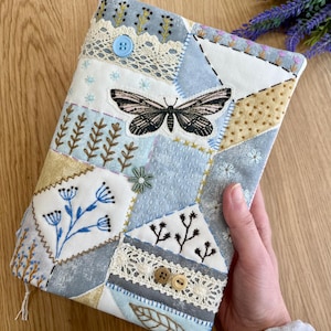 May include: A fabric book cover featuring a patchwork design in shades of blue, white, and tan. The cover is decorated with intricate embroidery of a moth, floral sprigs, and leaves, accented by white lace trim, a light blue button, and two wooden buttons.