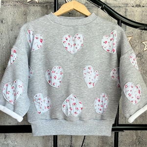 Puede incluir: Una sudadera gris con parches en forma de corazón. Cada corazón está relleno con un estampado floral de flores rosas y rojas sobre un fondo azul claro. Las mangas tienen puños blancos. La sudadera está colgada de una percha de madera.