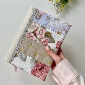 May include: A handmade fabric journal cover with a patchwork design featuring floral patterns in shades of pink, blue, and beige. The cover has a delicate lace trim and a pink ribbon.