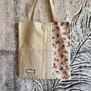 May include: A beige tote bag with a floral pattern on the right side and a lace trim. The left side is plain beige. The bag has beige handles and a small label that reads "IVA NOVA".