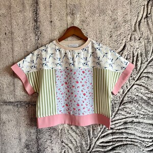 Puede incluir: Top de manga corta con diseño patchwork. El top presenta un cuello color crema, mangas rosas y un dobladillo inferior. Tiene un estampado floral, rayas verticales y un patrón de grúa.