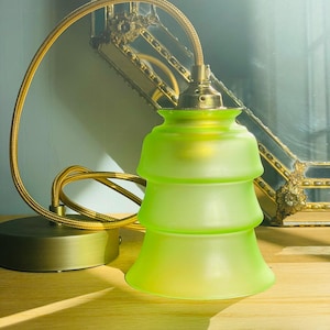 Könnte beinhalten: Eine Pendelleuchte mit einem abgestuften, mattierten lindgrünen Glasschirm. Die Lampe verfügt über eine messingfarbene Fassung und einen passenden runden Sockel, die durch ein flexibles, goldfarbenes Textilkabel verbunden sind.