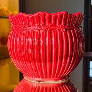 Puede incluir: Una maceta de cerámica roja vibrante con un diseño acanalado y un borde festoneado. La maceta presenta una guirnalda floral alrededor del borde superior. El acabado brillante refleja el entorno, creando un efecto visual llamativo.