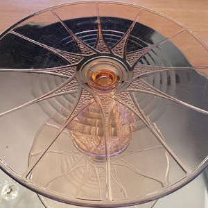 May include: A pink glass cake stand with a starburst design on the top tier. The stand has a detailed pattern and a central, raised circular base. The glass has a reflective quality, and the stand is on a reflective surface.