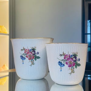 Op de afbeelding: Twee witte keramische potten met een bloemenmotief en een gouden rand. De potten hebben een geweven patroon en bevatten roze, blauwe en gele bloemen.
