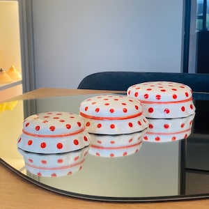 Op de afbeelding: Drie witte keramische sieradenboxen met rode stippen. De dozen zijn in een rij op een reflecterend oppervlak gerangschikt.