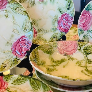 Könnte beinhalten: Ein Set antiker Keramikteller und -schalen mit floralem Design. Die Teller und Schalen sind mit rosa Rosen und grünen Blättern auf cremefarbenem Hintergrund verziert.