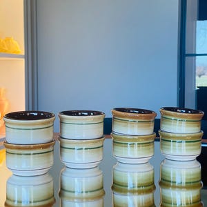 May include: Set of eight brown, green, and white ceramic cups with a striped pattern. The cups are arranged in two rows of four on a mirrored surface.