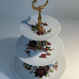Könnte beinhalten: Dreistöckiger Kuchenständer aus weißem Porzellan mit goldenem Metallgriff und Blumenmuster.