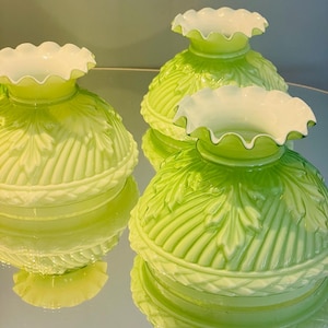 May include: Three green glass lamp shades with a ruffled white rim. The shades have a leaf pattern and are reflected on a mirrored surface. The shades are a light green color.