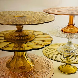 May include: A set of four amber glass cake stands with intricate patterns. The stands are stacked on top of each other, with the largest stand at the bottom and the smallest stand at the top.