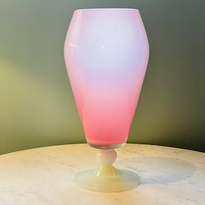 Puede incluir: Un jarrón de cristal alto con un cuerpo cónico que transiciona de un blanco suave en la parte superior a un tono rosa vibrante en el centro. El recipiente se apoya sobre un tallo corto y redondeado y una base circular ancha con acabado opalescente.