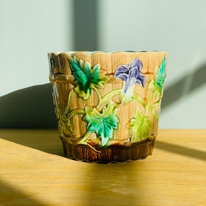 May include: A ceramic planter with a tan, wood-grain textured exterior featuring raised, glossy green leaves and a single purple morning glory flower. The base of the pot is a darker brown, and the top rim has a scalloped, wavy edge design.